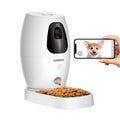 A white automatic pet feeder with a bowl of kibble on a clean white background. A human hand is holding a smartphone showing a dog photo, demonstrating the app control feature.