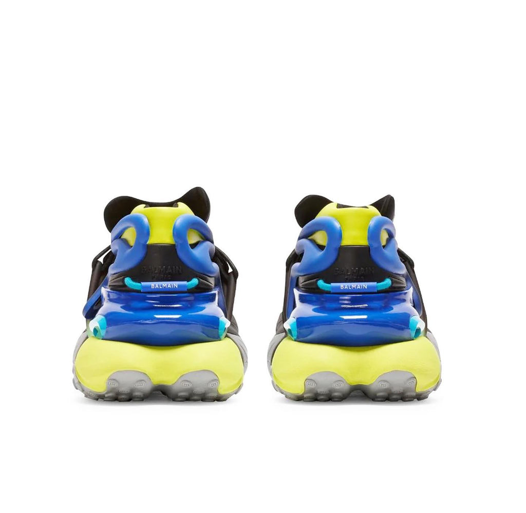 A pair of chunky sneakers shown from the back, featuring a bright neon yellow and blue design with black details and gray soles, set against a plain white background.