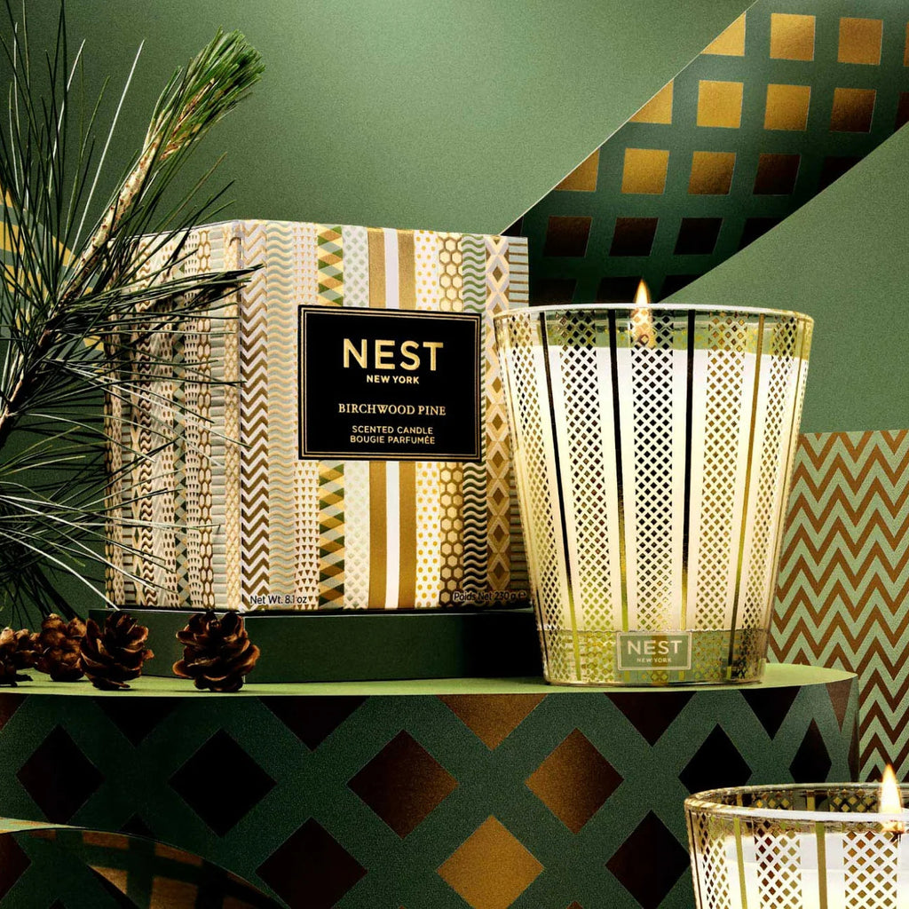 A Nest New York Birchwood Pine scented candle in a gold-toned metal cage holder, shown next to its patterned box on a green table with pine branches and decorative geometric background.