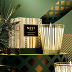 A Nest New York Birchwood Pine scented candle in a gold-toned metal cage holder, shown next to its patterned box on a green table with pine branches and decorative geometric background.