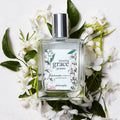 Perfume bottle labeled 'amazing grace jasmine' by philosophy surrounded by white flowers and green leaves on a light background.