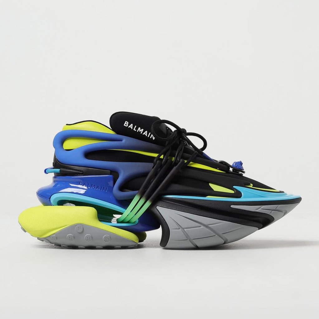 A pair of Balmain sneakers featuring a stacked, sculptural design in black, blue, gray, and neon yellow-green, with black laces and the Balmain logo visible on the tongue.