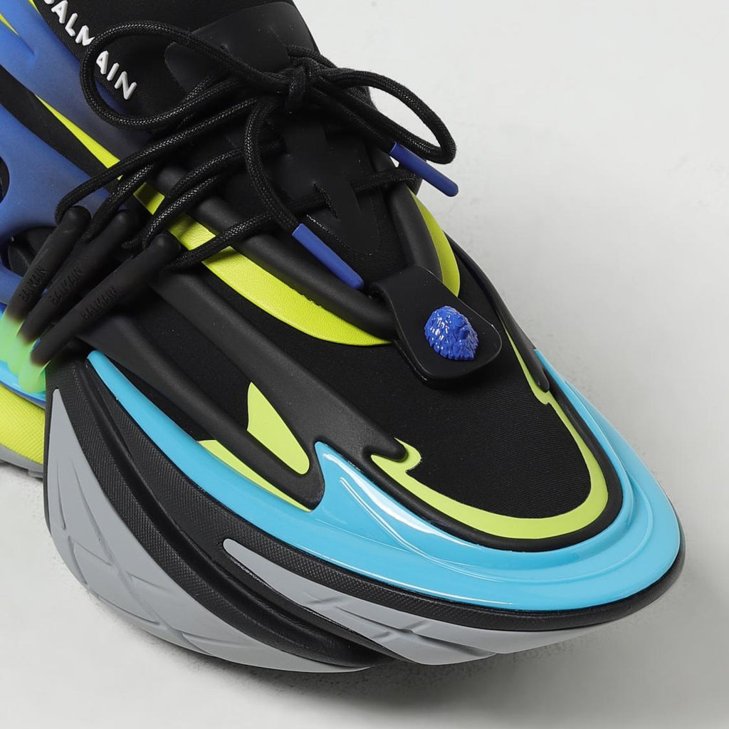 A close-up of a Balmain sneaker featuring a black upper with bright neon yellow-green accents, a light blue midsole, and a gray outsole.