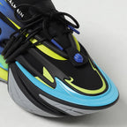 A close-up of a Balmain sneaker featuring a black upper with bright neon yellow-green accents, a light blue midsole, and a gray outsole.