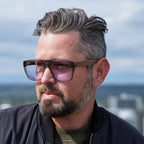 A man with styled gray hair and a beard wearing dark brown square sunglasses with pink-tinted lenses, photographed outdoors with a blurred sky and city background.