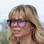 A close-up portrait of a woman with blonde hair wearing oversized square sunglasses with dark brown frames and pink-tinted lenses.